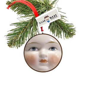 May include: A round, white ceramic ornament with a face, blue eyes, and rosy cheeks. The ornament is trimmed with a brown border and hangs from a red ribbon. A tag with the text "SILVA WARE" is attached to the ornament.