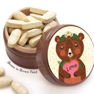 May include: A brown wooden pill box with a lid featuring a bear holding a pink heart with the word "Love". The box is filled with oblong, light-colored pills. The text "Shown in Brown Paint" is also visible.