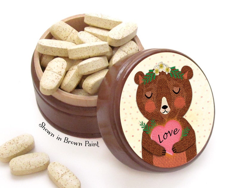 May include: A brown wooden pill box with a lid featuring a bear holding a pink heart with the word "Love". The box is filled with oblong, light-colored pills. The text "Shown in Brown Paint" is also visible.
