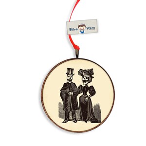 May include: Round wooden ornament featuring a black and white illustration of a skeleton couple in formal attire. The ornament has a light beige background and a dark brown wooden frame. A red ribbon is attached for hanging. The tag reads "Silva Ware".