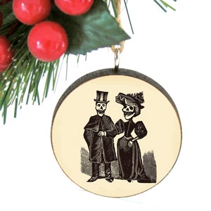 May include: A round wooden ornament featuring a black and white illustration of a skeleton couple in vintage clothing. The ornament is hanging with a twine loop, with red berries and green pine needles in the background.