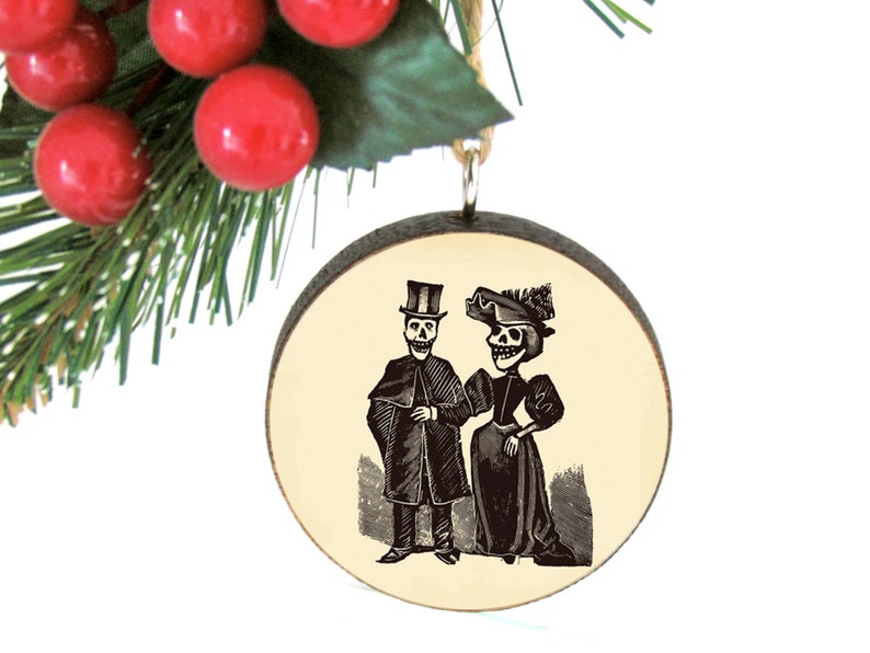 May include: A round wooden ornament featuring a black and white illustration of a skeleton couple in vintage clothing. The ornament is hanging with a twine loop, with red berries and green pine needles in the background.