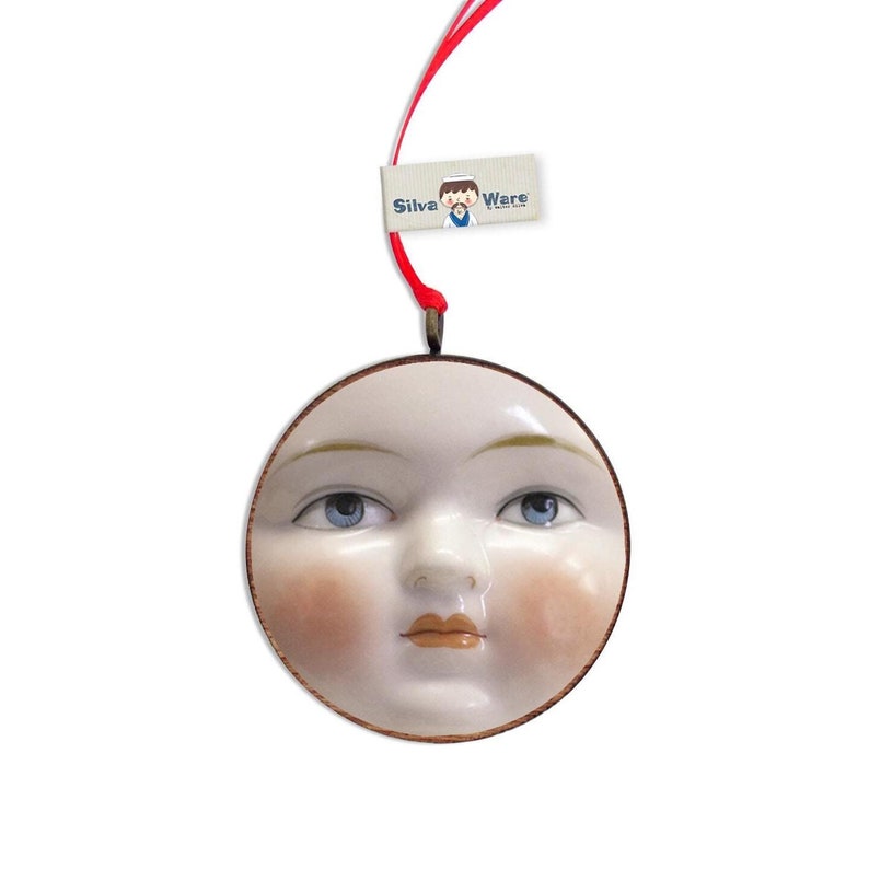 May include: A round, off-white ornament with a painted face featuring blue eyes, rosy cheeks, and a small, closed mouth. The ornament has a brown border and is suspended by a red ribbon. A tag that reads "Silva Ware" is attached.