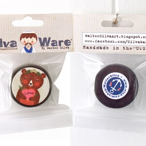 May include: Two round, dark brown decorative items in clear plastic bags. One features a bear holding a heart with the word "Love." The other has a white sticker with an anchor design. The packaging reads "Silva Ware" and "Handmade in the U.S.A."