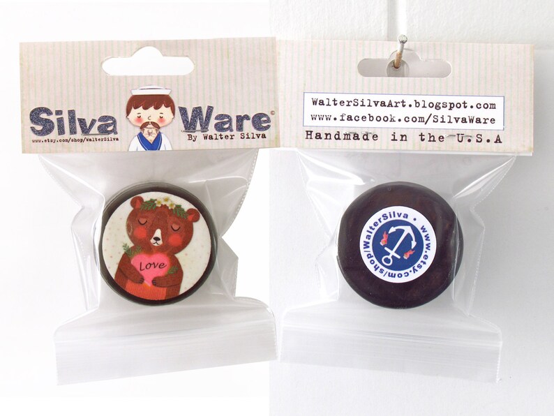 May include: Two round, dark brown decorative items in clear plastic bags. One features a bear holding a heart with the word "Love." The other has a white sticker with an anchor design. The packaging reads "Silva Ware" and "Handmade in the U.S.A."