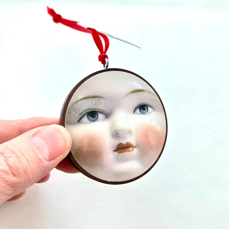 May include: A round ornament featuring a painted face with blue eyes, rosy cheeks, and a brown border. A red ribbon and needle are attached for hanging. The ornament is held by a hand against a white background.