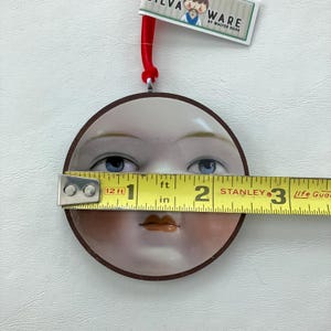 May include: A round ornament featuring a painted face with blue eyes and a brown border. The ornament measures approximately 2.5 inches in diameter. A red ribbon is attached for hanging. The tag reads "SILVA WARE by WALTER SILVA."