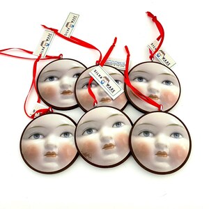May include: Six round ceramic ornaments with painted faces, each featuring blue eyes, rosy cheeks, and a brown border. Red ribbons are attached for hanging. Each ornament has a tag that reads "SILVA WARE".