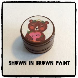 May include: A brown, round container with a lid featuring a cartoon bear holding a pink heart with the word "Love." The bear wears a floral crown. The text "SHOWN IN BROWN PAINT" is below the container.