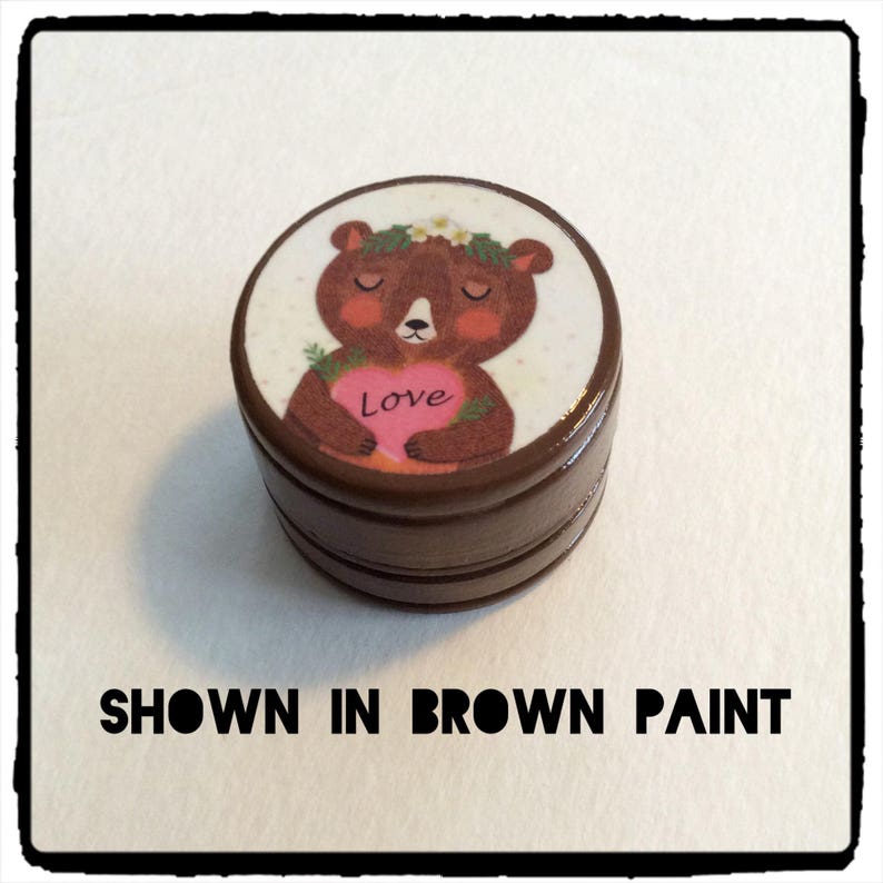 May include: A brown, round container with a lid featuring a cartoon bear holding a pink heart with the word "Love." The bear wears a floral crown. The text "SHOWN IN BROWN PAINT" is below the container.