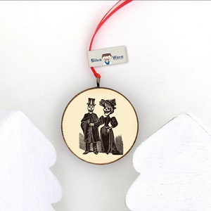 May include: Round wooden ornament featuring a vintage illustration of a skeleton couple in formal attire. The ornament has a red ribbon for hanging and a small tag that reads "Silva Ware". Two white, stylized Christmas trees are in the foreground.