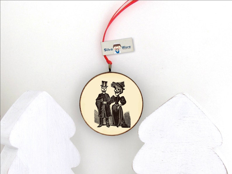 May include: Round wooden ornament featuring a vintage illustration of a skeleton couple in formal attire. The ornament has a red ribbon for hanging and a small tag that reads "Silva Ware". Two white, stylized Christmas trees are in the foreground.
