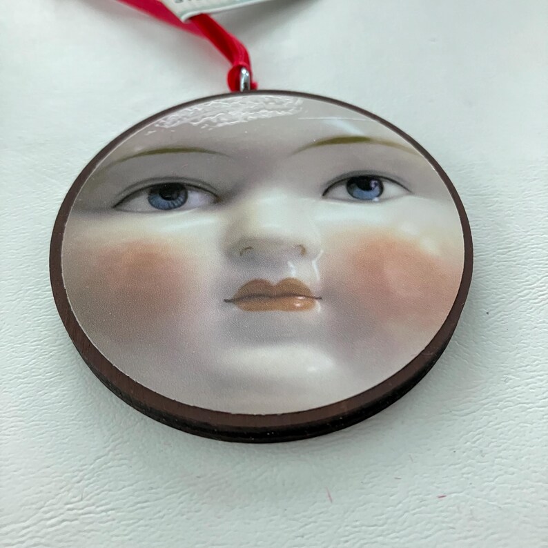 May include: A round ornament featuring a serene face with blue eyes, rosy cheeks, and a neutral lip color. The face is set against a white background, framed by a dark brown border. A red ribbon is attached for hanging.