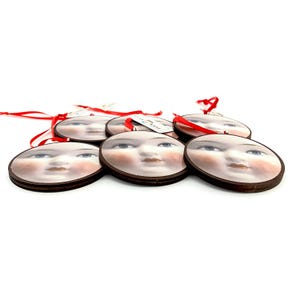 May include: A collection of six round ornaments featuring a vintage-style face design with blue eyes, rosy cheeks, and a neutral lip color. Each ornament has a dark brown wooden border and a red ribbon for hanging. The ornaments are on a white background.