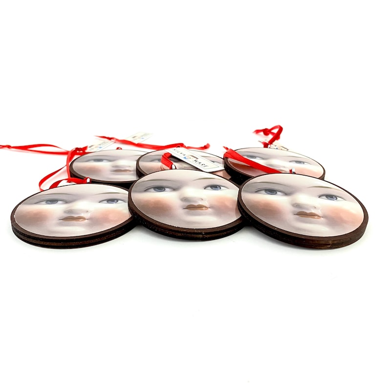 May include: A collection of six round ornaments featuring a vintage-style face design with blue eyes, rosy cheeks, and a neutral lip color. Each ornament has a dark brown wooden border and a red ribbon for hanging. The ornaments are on a white background.