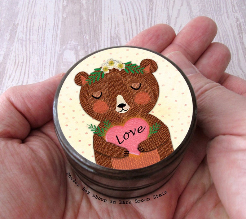 May include: A round, dark brown powder box with a lid featuring a cartoon bear holding a pink heart with the word "Love." The bear has a floral crown and is set against a dotted background. The text "Powder box shown in Dark Brown stain" is at the bottom.