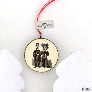 May include: Round, double-sided wooden ornament with a vintage illustration of a skeleton couple in formal attire. The ornament has a red ribbon for hanging and a small tag that reads "Silva Ware". The background is cream-colored.