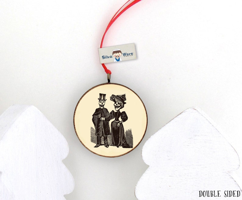 May include: Round, double-sided wooden ornament with a vintage illustration of a skeleton couple in formal attire. The ornament has a red ribbon for hanging and a small tag that reads "Silva Ware". The background is cream-colored.