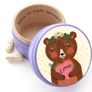 May include: A light purple round trinket box with a wooden interior and a lid featuring a cartoon bear holding a pink heart with the word "Love." The box is open, with the text "Shown in Light Lavender" inside. Several pills are scattered around the box.