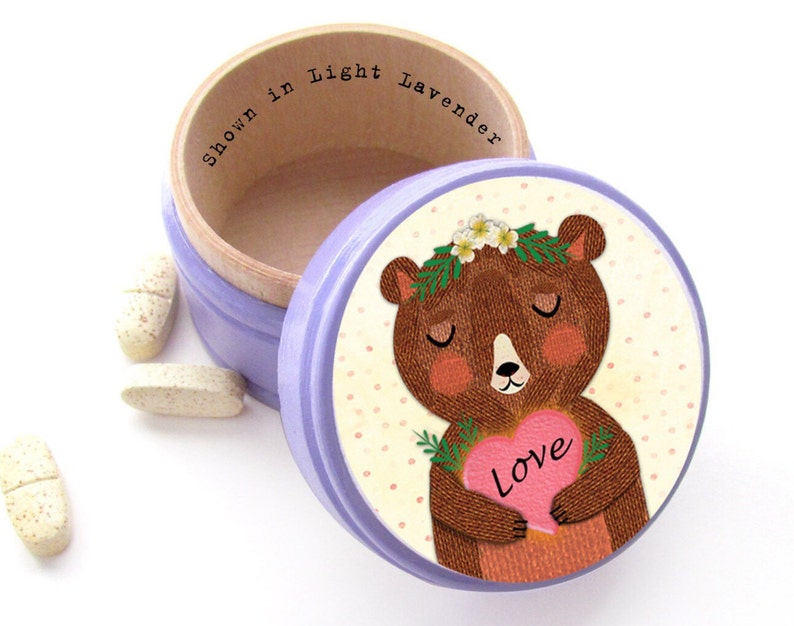 May include: A light purple round trinket box with a wooden interior and a lid featuring a cartoon bear holding a pink heart with the word "Love." The box is open, with the text "Shown in Light Lavender" inside. Several pills are scattered around the box.