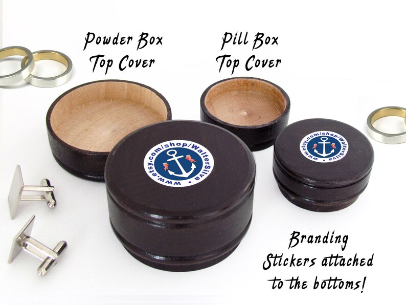 May include: A collection of small, dark brown wooden boxes, including a powder box and pill boxes, with light-colored interiors. The boxes have branding stickers with an anchor logo. Also shown are silver cufflinks and rings.