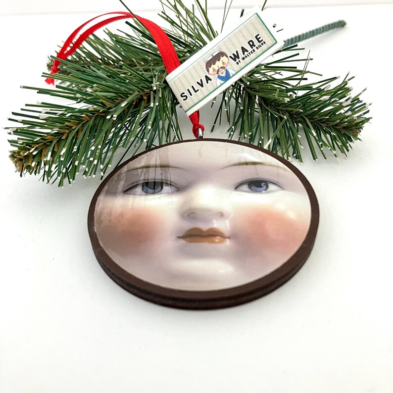 May include: A round Christmas ornament featuring a serene face with blue eyes and rosy cheeks. The ornament has a brown wooden frame and a red ribbon for hanging. The tag reads "SILVA WARE by WALTER SILVA".
