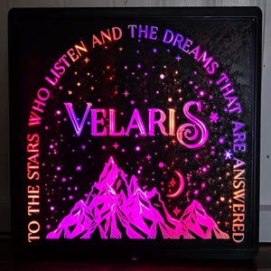 Może przedstawiać: Podświetlany znak w czarnej ramie z łańcuchem g&oacute;rskim, rosnącym księżycem i gwiazdami. Tekst brzmi "To the stars who listen and the dreams that are answered Velaris".