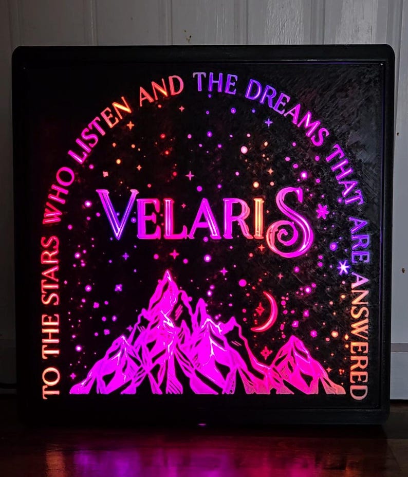 Może przedstawiać: Podświetlany znak w czarnej ramie z łańcuchem g&oacute;rskim, rosnącym księżycem i gwiazdami. Tekst brzmi "To the stars who listen and the dreams that are answered Velaris".
