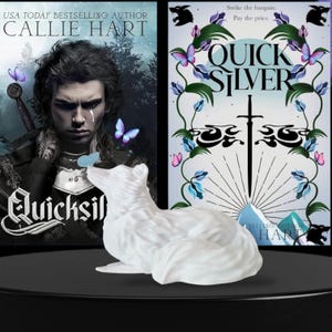 Puede incluir: Una figura de gato de cerámica blanca se encuentra frente a dos portadas de libros. Las portadas de los libros son para "Quicksilver" de Callie Hart. Las portadas tienen un tema de fantasía con mariposas y una espada.