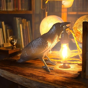Könnte beinhalten: Eine bronzefarbene Krähenskulptur, die eine Glühbirne hält, auf einer Holzoberfläche. Die Krähe ist mit strukturierten Federn detailliert und steht in der Nähe einer leuchtenden Glühbirne und zweier Globen. Die Szene wird von warmem Licht beleuchtet.
