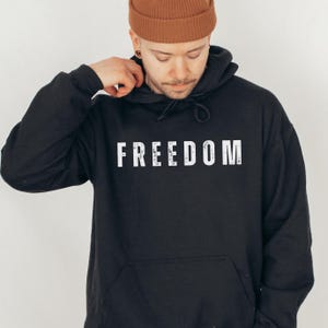 May include: Black hoodie with the word "FREEDOM" printed in white across the chest. The hoodie has a drawstring hood and a kangaroo pocket. The person in the image is wearing a brown beanie.