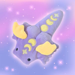 May include: A plush toy in the shape of a stylized creature, primarily lavender with yellow accents. The toy features embroidered crescent moons and stars, with two yellow wing-like shapes. Black button eyes complete the design.