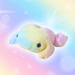 Könnte beinhalten: Ein Plüschtier in Walform mit einem weichen, mehrfarbigen Design. Der Wal hat einen Farbverlauf von Gelb zu Blau und Rosa, kleine schwarze Augen und flossenartige Anhänge. Ein Spielzeug für Kinder.