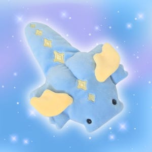 Puede incluir: Un peluche con forma de criatura azul con detalles amarillos. El juguete tiene orejas amarillas y patrones en forma de diamante. El fondo es un degradado de azul y morado con estrellas blancas.