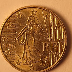 May include: A gold-coloured 1999 Irish 1 Euro coin featuring a woman holding a spear and a shield, with the inscription "ÉIRE" and twelve stars around the edge.