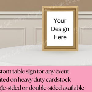 May include: A gold-framed sign with the text "Your Design Here" on a white background. The sign is on a white tablecloth. The bottom of the image has text that reads "Custom table sign for any event".