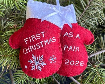 May include: A pair of red felt mittens joined together as a Christmas ornament with a white felt cuff and a white ribbon hanger. The embroidery reads FIRST CHRISTMAS AS A PAIR 2026, accompanied by a white stitched snowflake and a small star design.