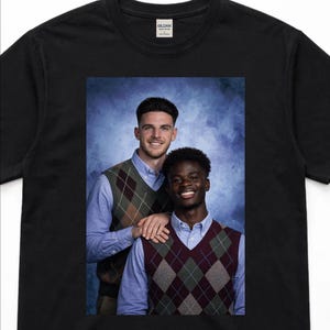 May include: Black t-shirt featuring a portrait of two people wearing argyle vests over blue button-down shirts. The background is a gradient of blue hues. The Gildan tag is visible on the shirt.