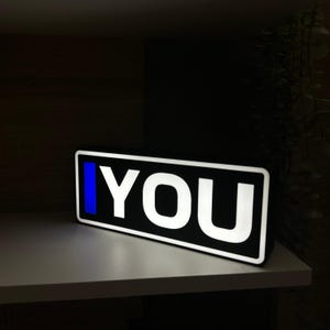 Può includere: Un cartello rettangolare in bianco e nero con la parola "YOU" in lettere bianche in grassetto. Una striscia blu verticale è sul lato sinistro. Il cartello è illuminato e poggia su una mensola bianca.