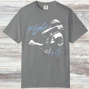 May include: Gray t-shirt featuring a white graphic of a basketball player in action, with the word "Flight" in light blue script. The player's signature is also in light blue. The shirt is made of a soft material.
