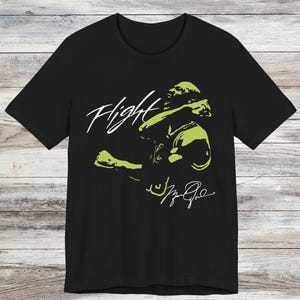 以下が含まれることがあります： 黒いTシャツ。ライムグリーンのグラフィックでバスケットボール選手がアクションを起こしている様子が描かれ、白い筆記体で「Flight」と書かれています。選手のサインが下にあります。