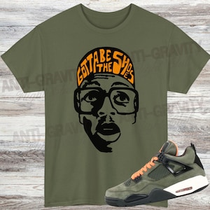 May include: An olive green t-shirt with a black and orange graphic of a person wearing glasses and the text "Gotta Be The Shoes." A pair of green, black, and orange trainers is displayed next to the shirt.