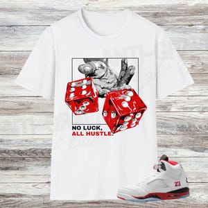 May include: White t-shirt with a graphic of two red dice and a hand, and the text "NO LUCK, ALL HUSTLE." A white and red sneaker with the number 23 is also visible. The shirt is displayed on a wooden background.