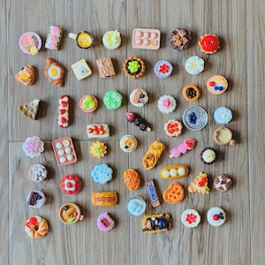 May include: A selection of miniature food charms, including pastries, fruits, and desserts. The charms are in various shapes and colours, such as a miniature pizza, a waffle, and a small pie. The charms are arranged on a wooden surface.