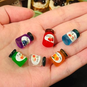 Op de afbeelding: Zes mini glazen potjes met kleurrijke etiketten en deksels. De potjes bevatten verschillende soorten jam, waaronder aardbei, pruim, vijg, abrikoos en watermeloen. De potjes zijn op een hand gerangschikt.