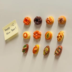 Puede incluir: Una colección de 12 figuritas de pan y pastelería en miniatura, coloridas y detalladas. Las figuritas están dispuestas en tres filas de cuatro sobre una superficie blanca. Una nota adhesiva amarilla con el texto "Happy New Year!" está en la esquina superior izquierda de la imagen.