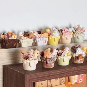 Op de afbeelding: Een verzameling mini-ijsbekers met verschillende toppings, waaronder chocolade, sprinkels en gummyberen. De ijsbekers worden gepresenteerd in kleine, witte, keramische kommen met een druppeleffect.
