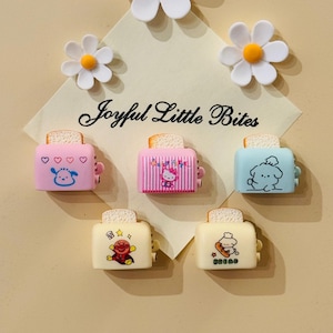 Op de afbeelding: Zes mini-toasters met verschillende cartoonfiguren erop. De toasters zijn roze, blauw en wit. De tekst "Joyful Little Bites" staat op de achtergrond.