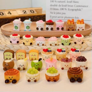 May include: A collection of miniature food charms, including cakes, pastries, bread, and drinks. The charms are made of polymer clay and are painted in various colors, including pink, green, brown, and white. The charms are arranged on a woven wicker surface.