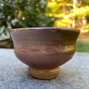 May include: A handmade ceramic bowl with a pedestal base. The bowl is a warm brown color with a textured surface and a leaf-like design. The base is a lighter shade of brown. The bowl is sitting on a gray surface.